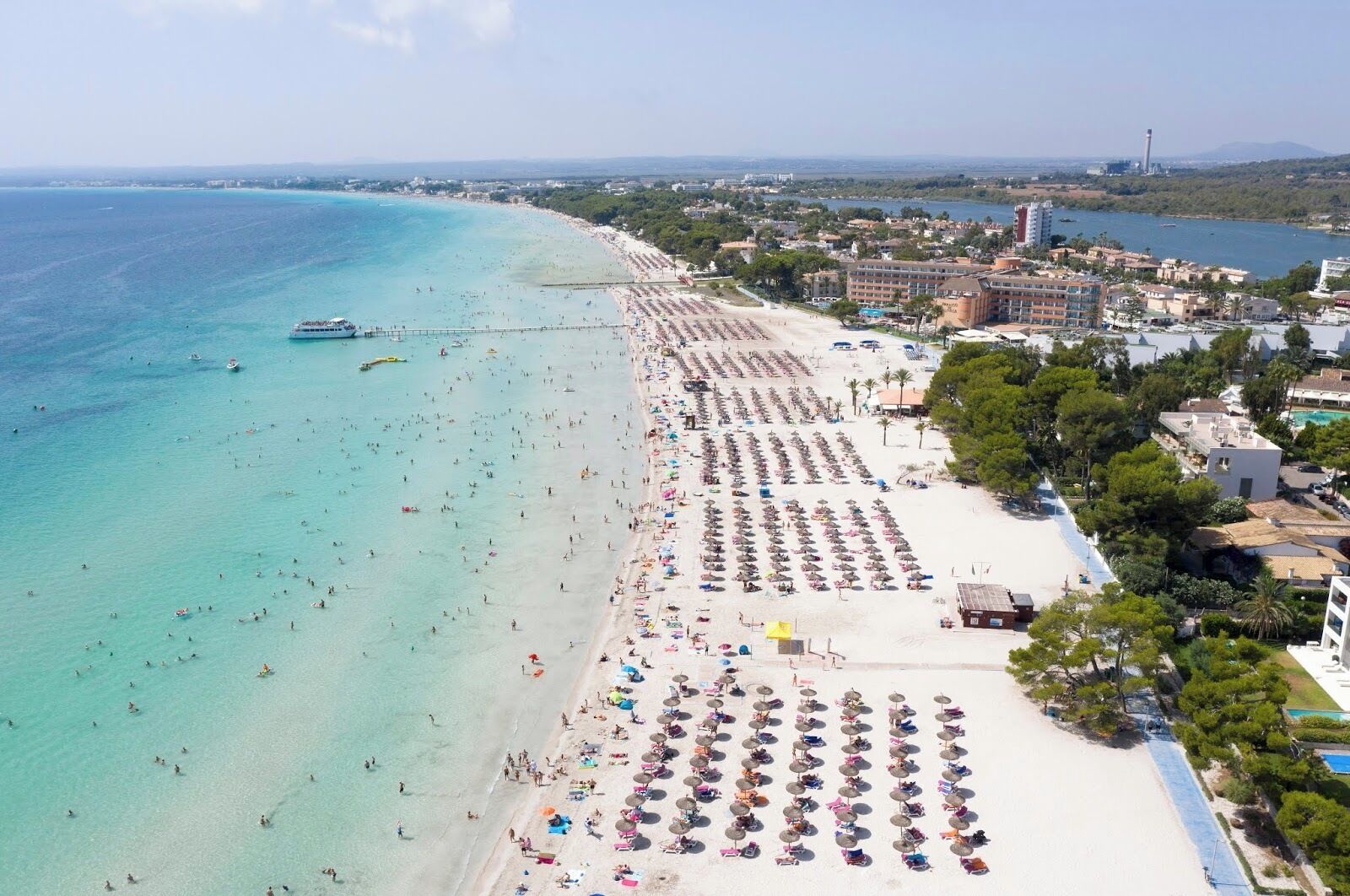 Una playa con hileras de sombrillas y sillas de baño, un mar azul cristalino y edificios al fondo.