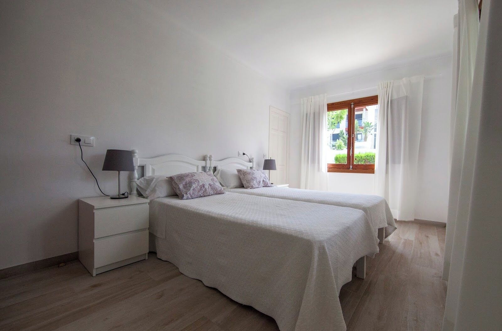 Un dormitorio con dos camas, una mesita de noche con lámpara, una ventana con cortinas y vistas al exterior.