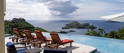 An infinity pool with lounge chairs and a table, overlooking a tropical landscape with a view of the ocean and islands.