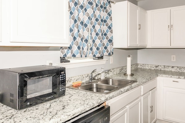 A kitchen with white cabinets, a stainless steel sink, a microwave, and a window with patterned curtains.