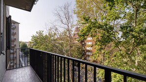 Un balcón con barandilla negra, con vistas a una calle con edificios y árboles.