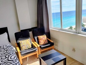 Ein Zimmer mit einem Bett, zwei Sesseln mit gemusterten Kissen, einem kleinen Tisch und Meerblick durch ein Fenster.