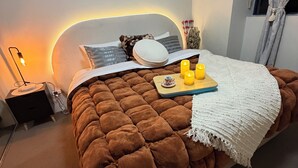 A bedroom with a tufted brown bedspread, a bedside table with a lamp, and a wall-mounted light fixture.