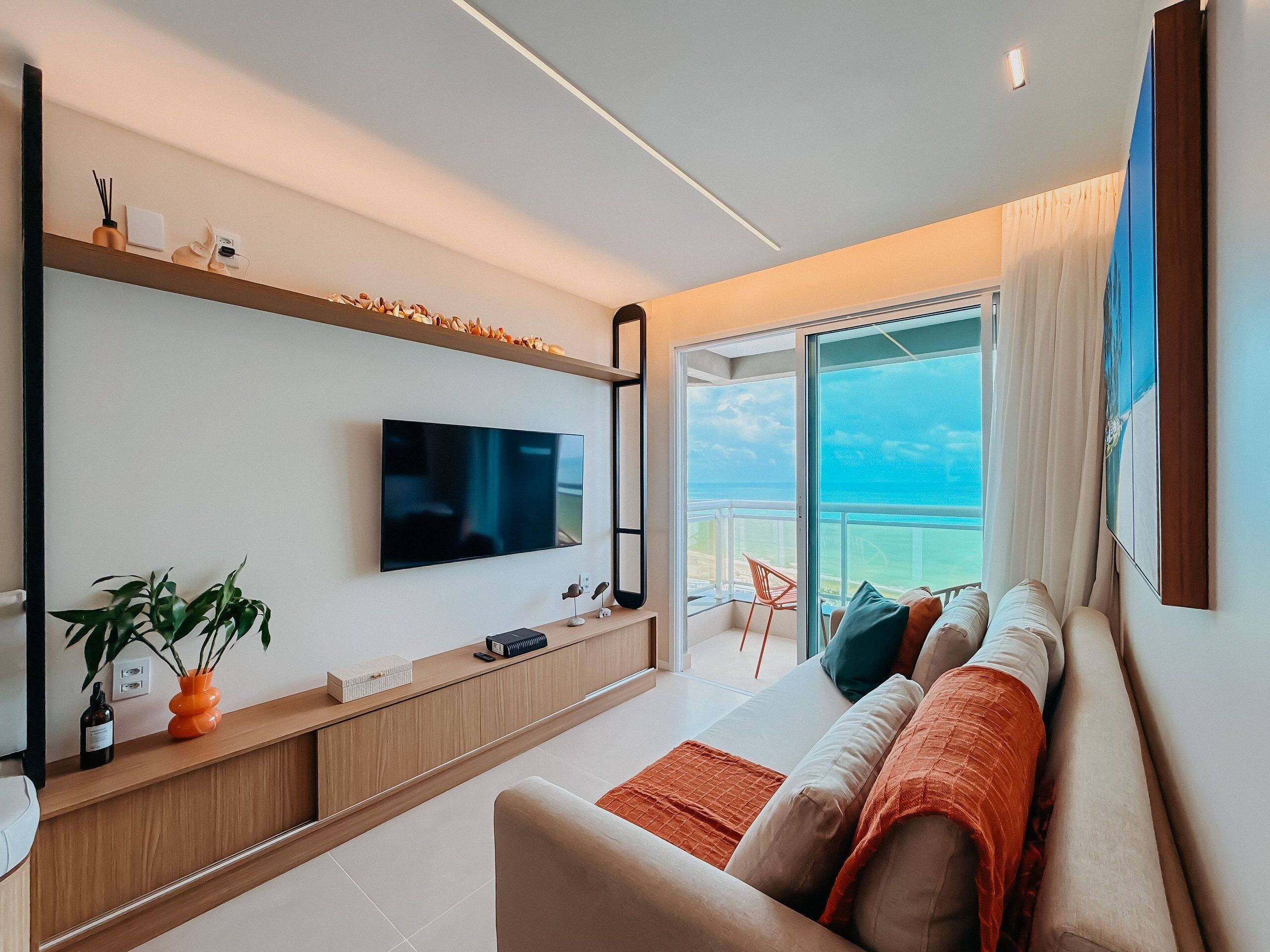 Uma sala de estar moderna com uma televisão de tela plana, uma mesa de console de madeira, um sofá com almofadas e vista para o oceano através de uma grande janela.