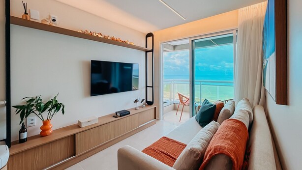 A modern living room with a flat-screen TV, a wooden console table, a sofa with cushions, and a view of the ocean through a large window. - Ahead Ponta Negra by Almare (Natal)