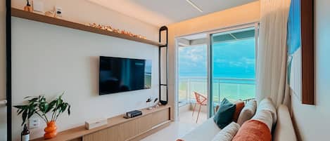 A modern living room with a flat-screen TV, a wooden console table, a sofa with cushions, and a view of the ocean through a large window.