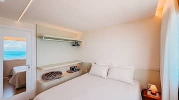 A hotel room with a bed, a bedside table with a lamp, a mirror, a shelf with a coat, and a window with a view of the sea.