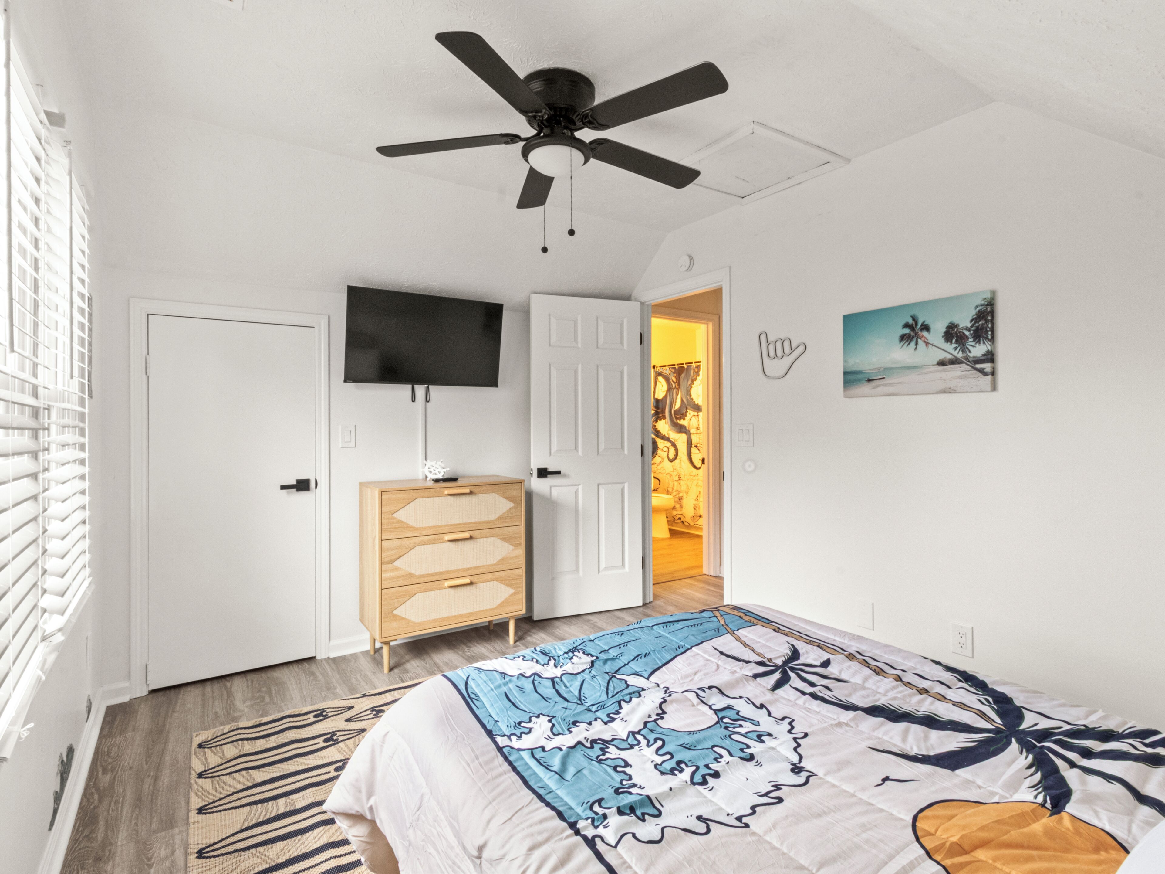 A bedroom with a bed, a ceiling fan, a TV, a dresser, and a door leading to a bathroom.