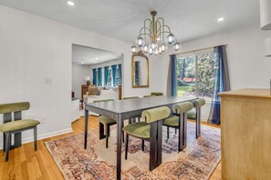 A dining area with a long table, multiple chairs, a chandelier, and a view of the outdoors through large windows. - Buckhead Hideaway ~ 4BR, Fire Pit, Big Deck! (Atlanta)