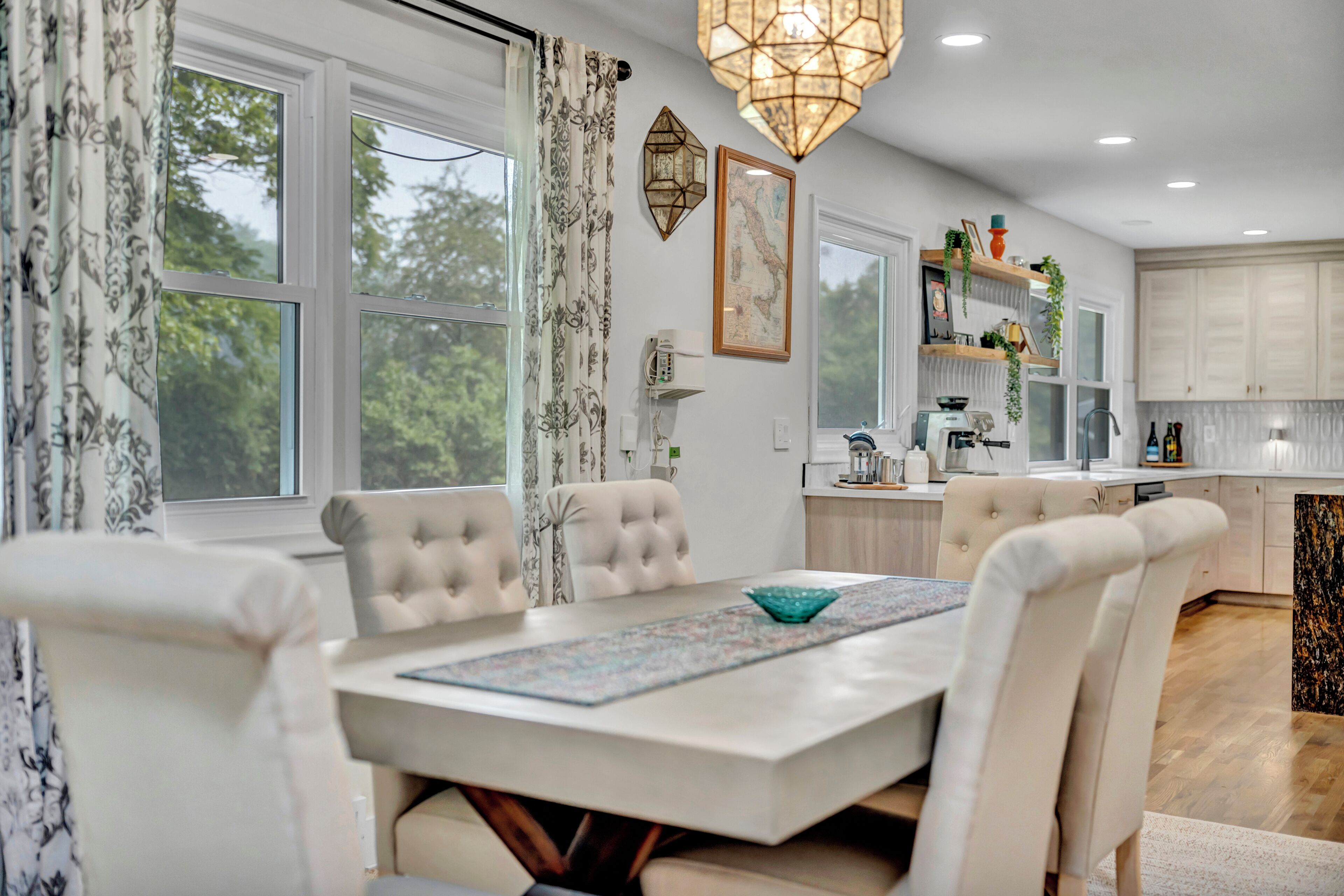 Une cuisine moderne avec des meubles blancs, un coin repas doté d’une table et de chaises blanches, et une grande fenêtre agrémentée de rideaux.