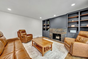 A modern living room with leather sofas, a wooden coffee table, a stone fireplace, and built-in shelving. - The Spartan Ranch (East Lansing)