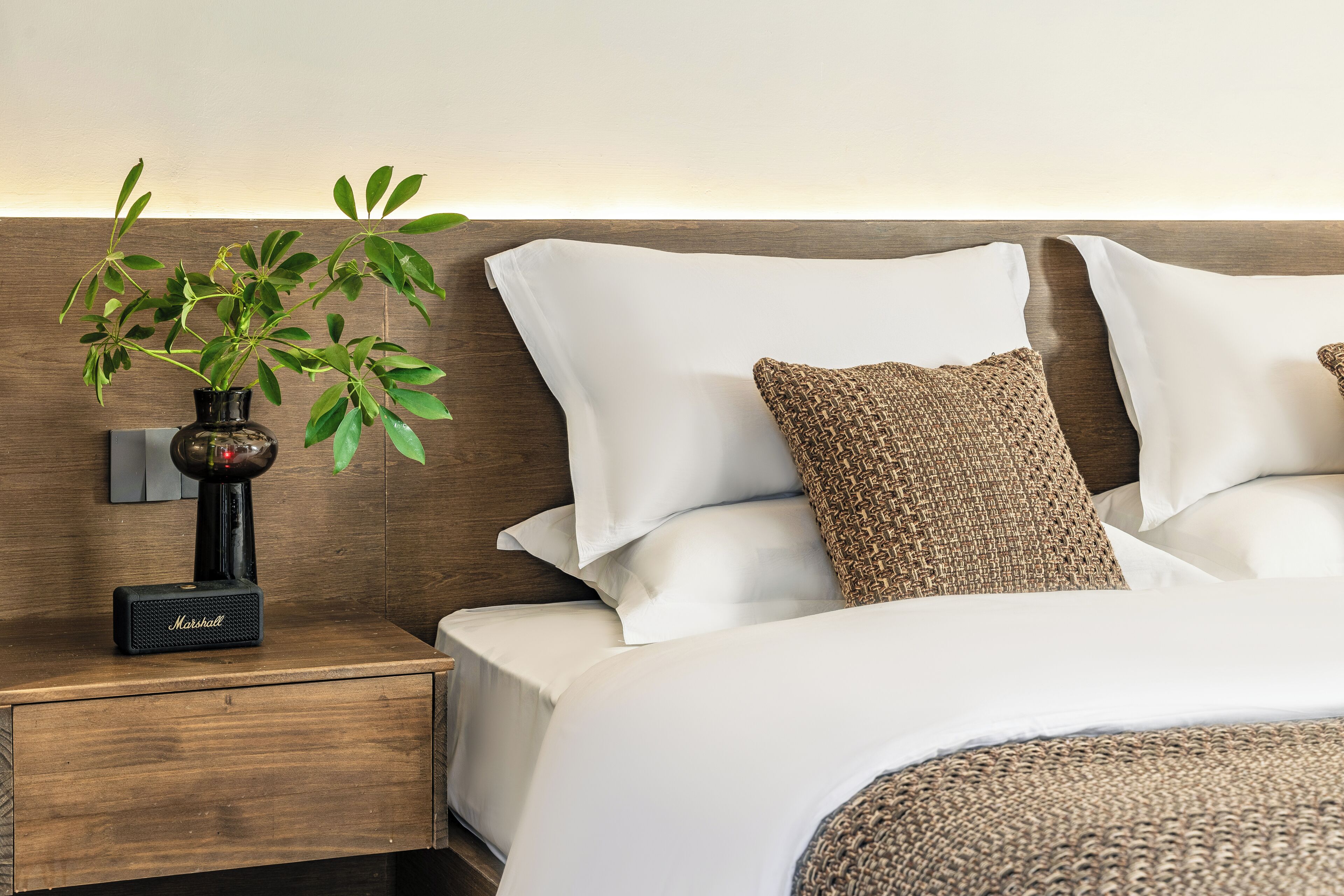 Un lit bien fait, avec du linge de lit blanc et un coussin marron tissé, une petite plante dans un vase noir posée sur une table de chevet en bois, et un haut-parleur Marshall.