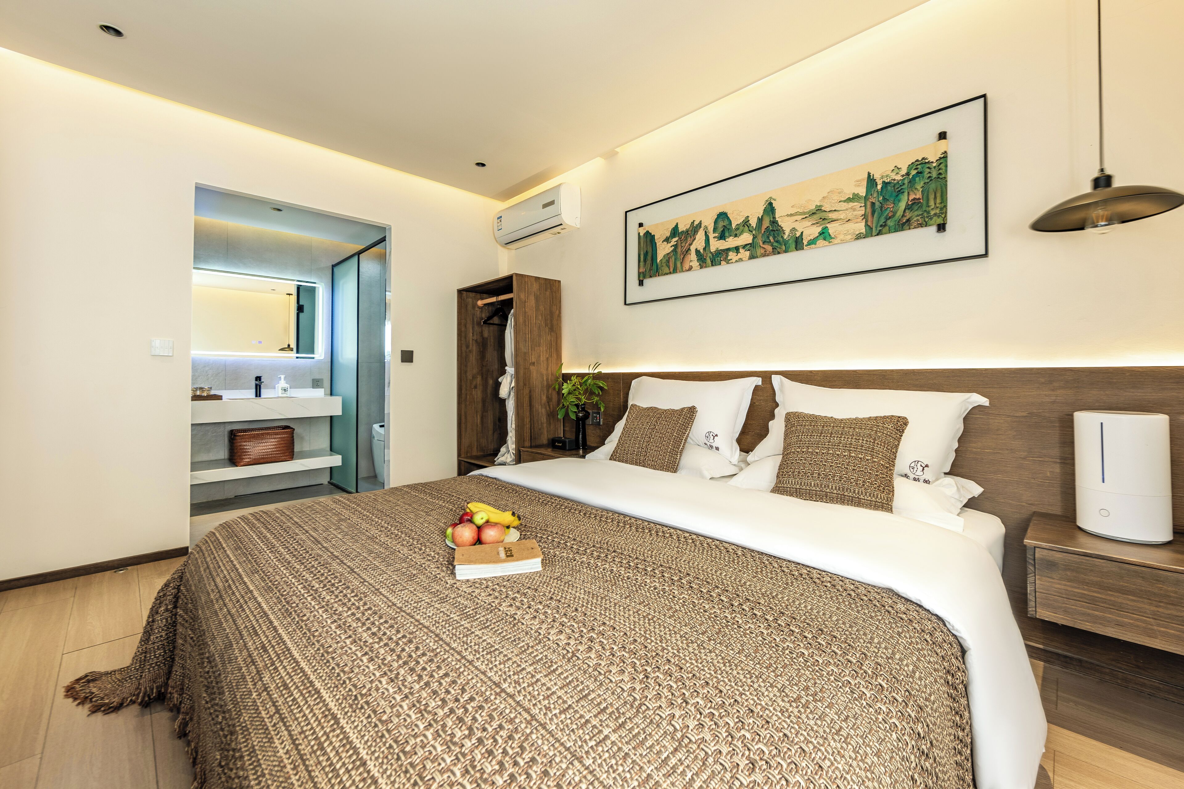 A modern hotel room with a large bed, a bedside table, a nightstand, a wall-mounted air conditioner, a framed painting, and a bathroom visible through an open door.