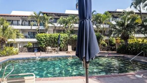 A swimming pool with a clear blue water surface, surrounded by palm trees and a building with balconies. - New Listing, Casa De Emdeko, Ocean views,AC (Kailua Kona)