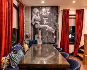Ein Zimmer mit einem großen Tisch, blauen Stühlen, roten Vorhängen und einem Wandgemälde, das eine Gitarre spielende Person zeigt.
