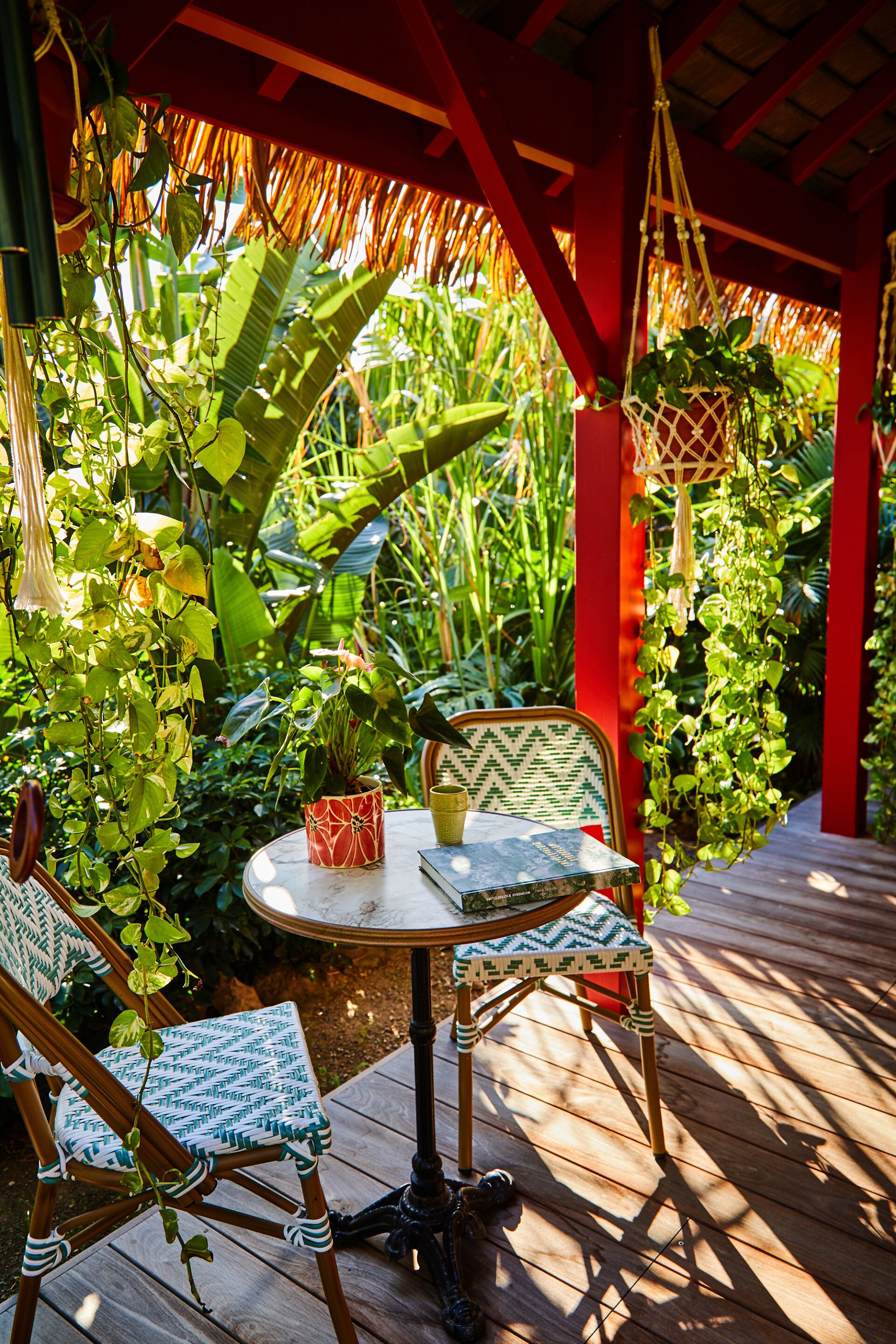 Un área de estar al aire libre con una mesa redonda y dos sillas, rodeada de abundante vegetación y una estructura roja.