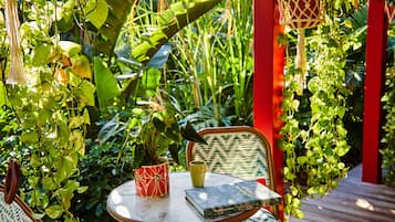 An outdoor seating area with a round table and two chairs, surrounded by lush greenery and a red structure.