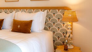 A neatly made bed with white bedding and brown pillows, a bedside lamp, and two framed botanical prints on the wall.
