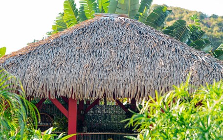 A thatched-roof structure with a wooden table and chairs, surrounded by lush greenery and a stone pathway.