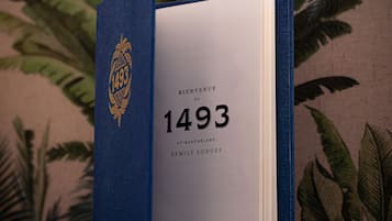Um livro intitulado 'BIENVENUE AU 1493' com um emblema decorativo na capa, colocado sobre uma superfície com padrão.