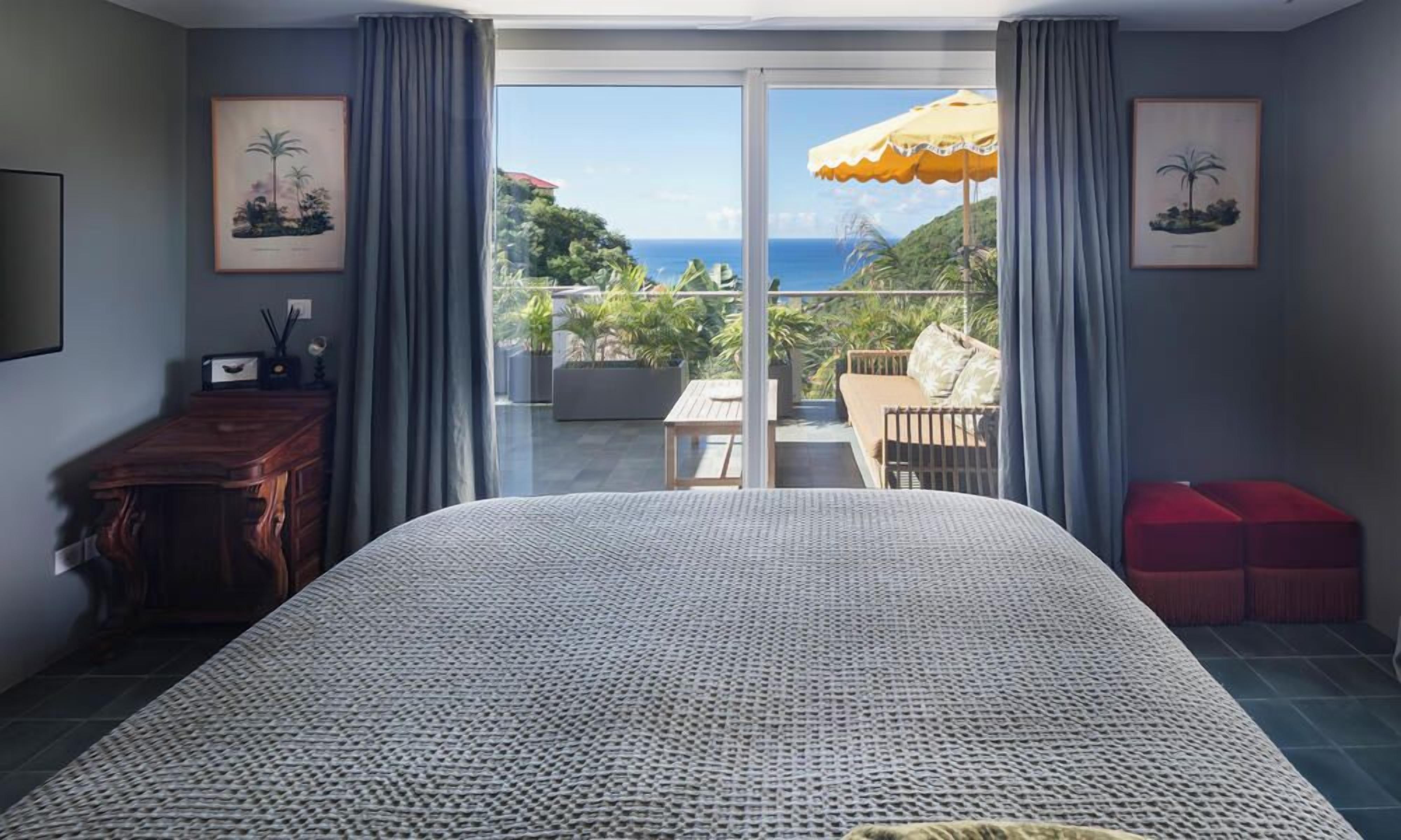 Une chambre avec un grand lit, une commode en bois, un pouf rouge et un balcon avec vue sur l’océan.
