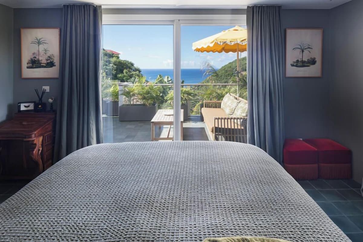 A bedroom with a large bed, a wooden dresser, a red ottoman, and a balcony with a view of the ocean.