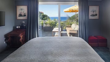 A bedroom with a large bed, a wooden dresser, a red ottoman, and a balcony with a view of the ocean.