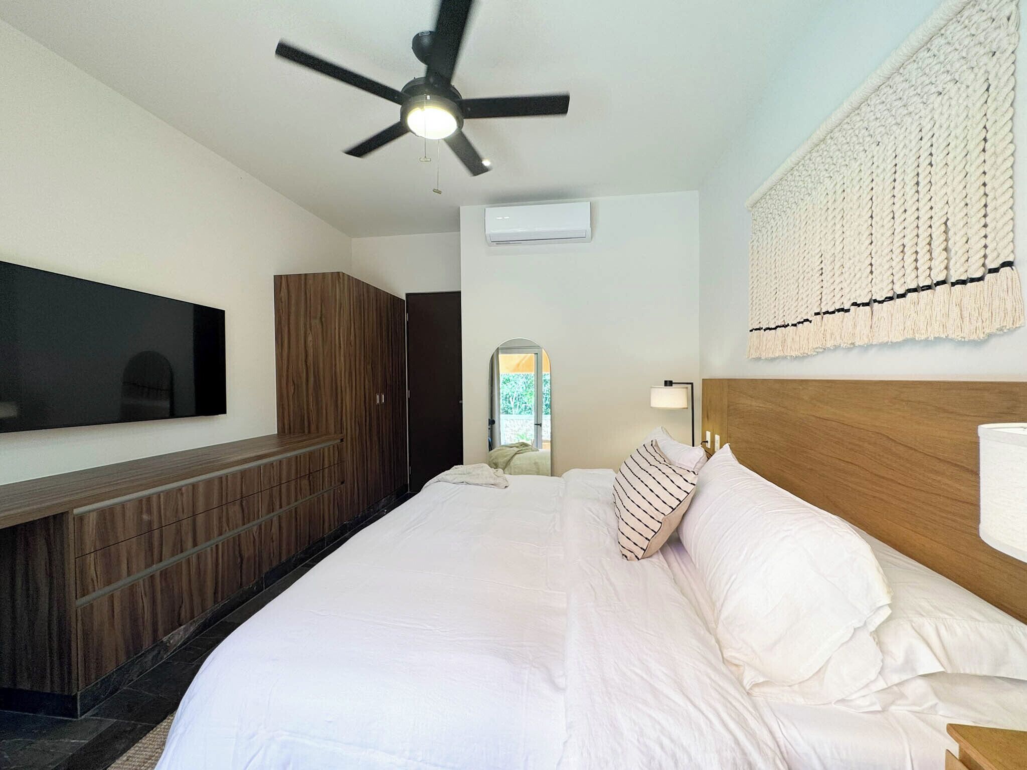 Une chambre d’hôtel comprenant un lit, un ventilateur de plafond, un téléviseur à écran plat, une armoire en bois, un climatiseur mural et un élément décoratif fixé au mur.