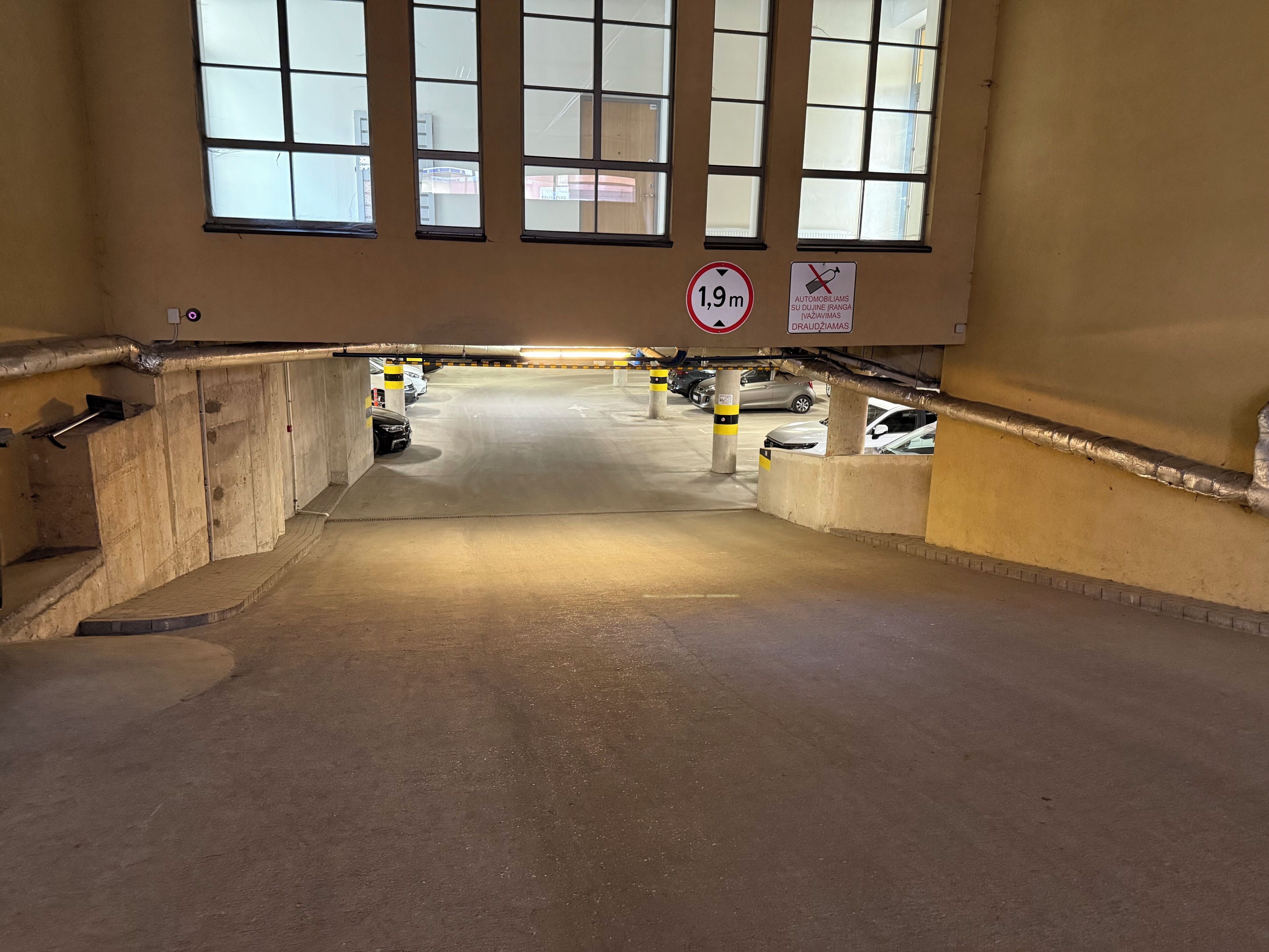 An underground parking garage with a 1.9m height limit sign, concrete pillars, and a clear view of parked cars.