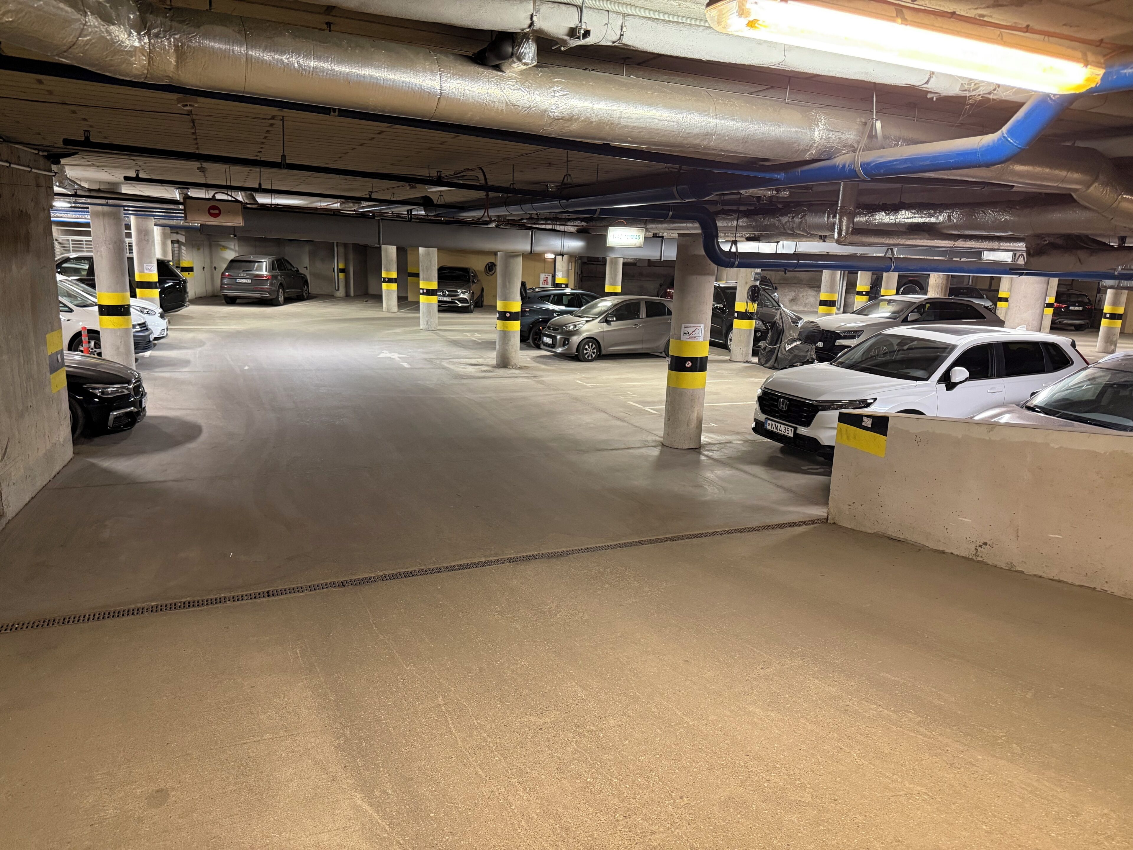 Un aparcamiento subterráneo de varios niveles con varios coches estacionados, pilares de hormigón con franjas amarillas y conductos de ventilación visibles.