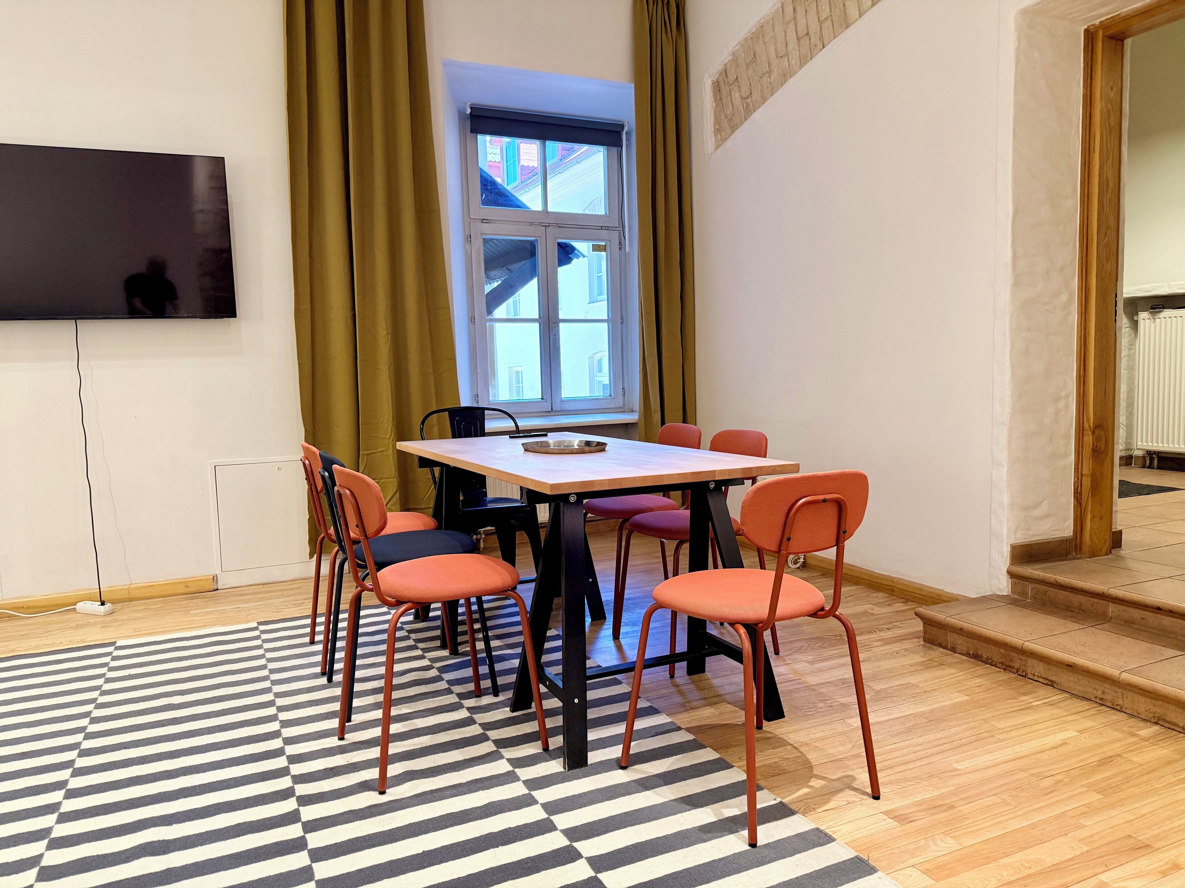 Ein Zimmer mit Esstisch, Stühlen, einem an der Wand befestigten Fernseher, einem Fenster mit Vorhängen und einem gemusterten Teppich.