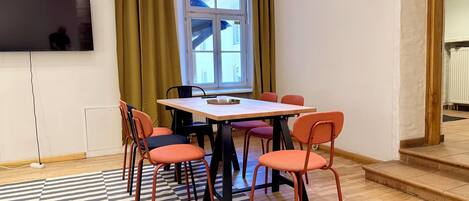 A room with a dining table, chairs, a TV mounted on the wall, a window with curtains, and a patterned rug.