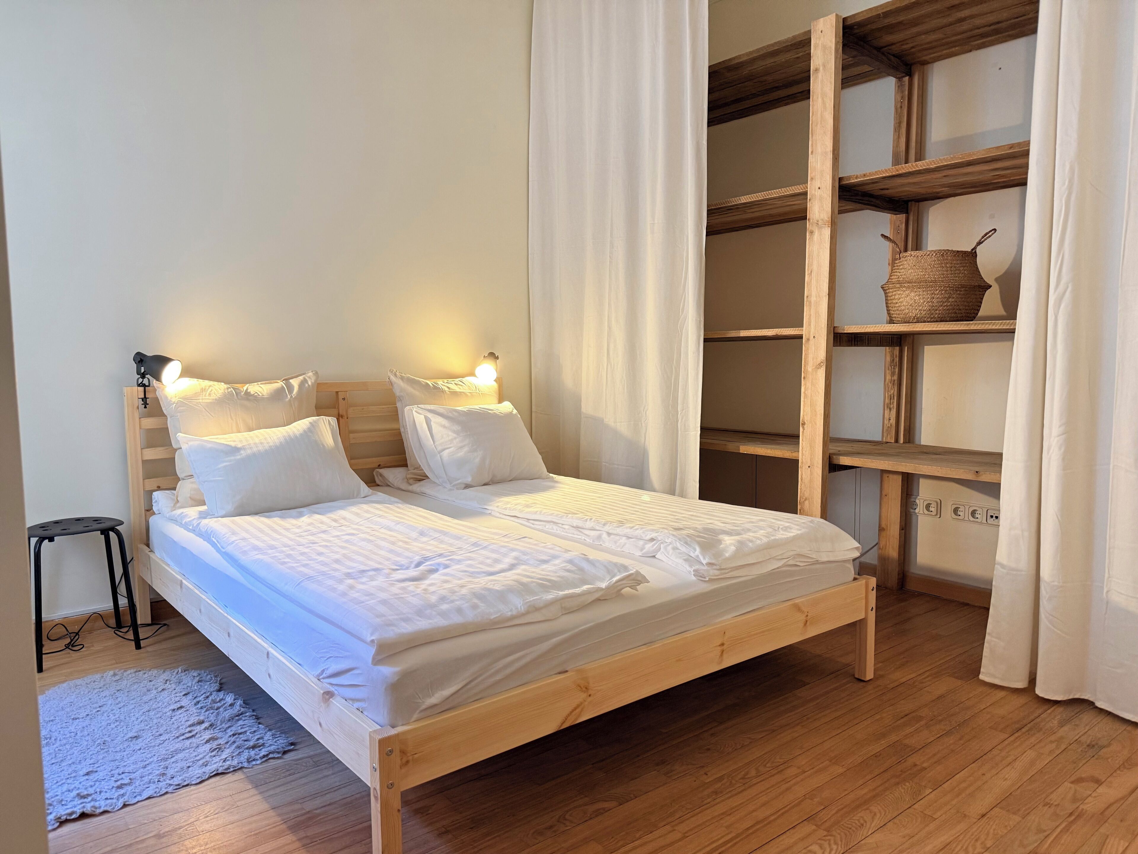 A wooden bed with white bedding, a bedside lamp, a small stool, and a woven basket on a shelf.