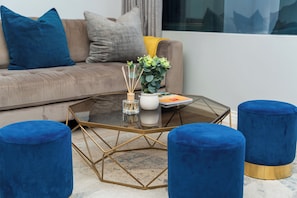 A modern living room with a glass coffee table, blue upholstered ottomans, a beige sofa with blue and gray cushions, and a potted plant with green foliage. - Luxury CBD Retreat | Stunning Mountain Views (Cape Town)
