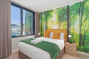 A hotel room with a large window, a bed with a green blanket, a headboard with a forest mural, and a nightstand with a lamp. - Luxury CBD Retreat | Stunning Mountain Views (Cape Town)