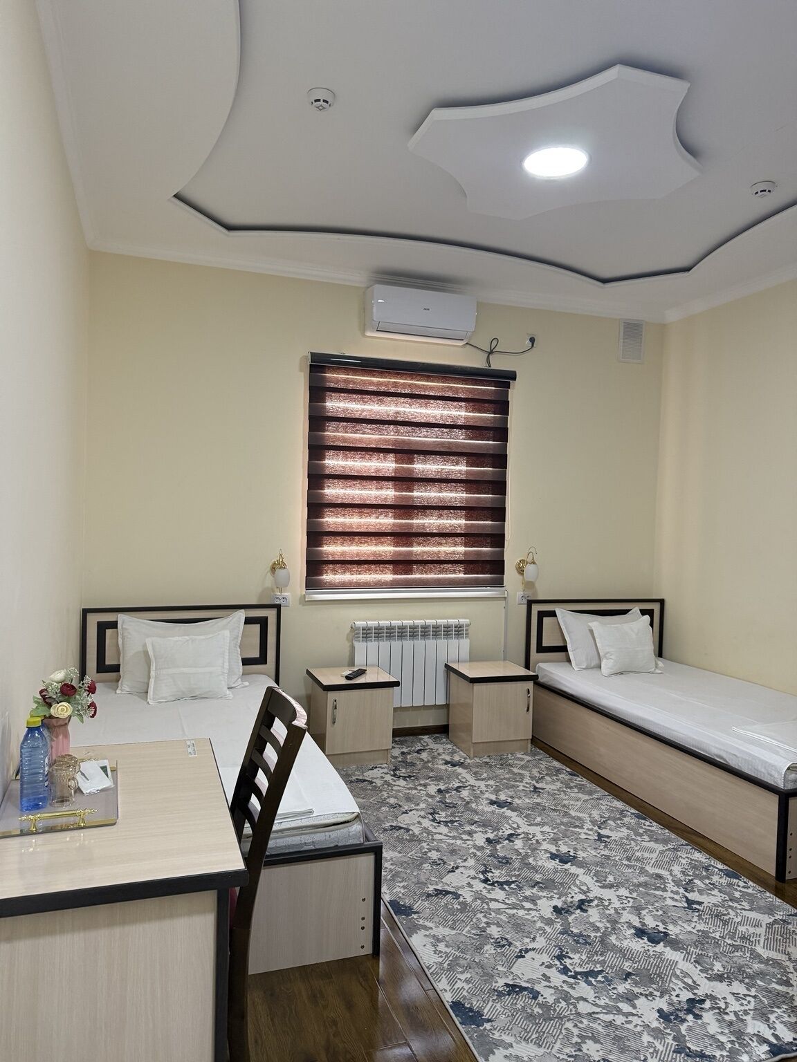 Une chambre d’hôtel avec deux lits simples, un bureau avec une chaise, une petite table, un radiateur et un tapis à motifs.