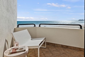 Ein Balkon mit einem weißen Stuhl, einem kleinen Tisch und einem Buch. Blick auf einen Strand mit dem Meer und einer entfernten Insel.