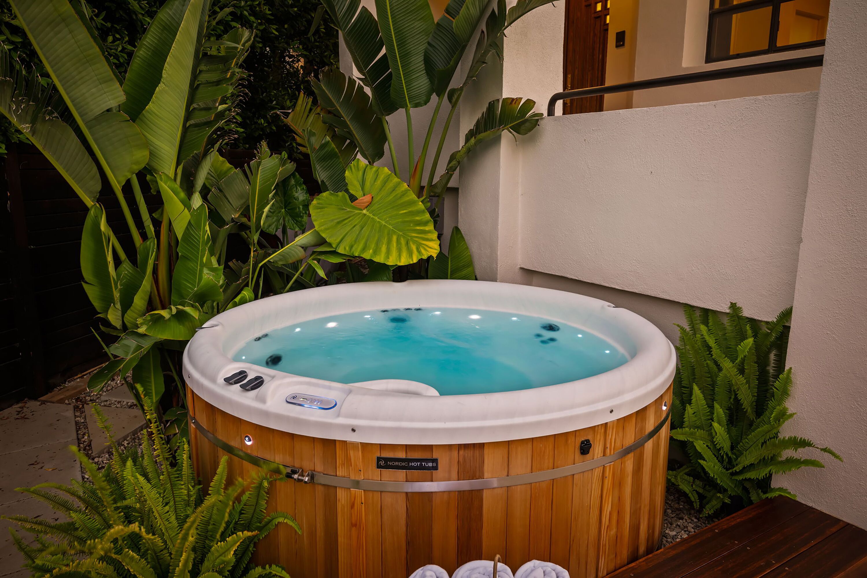 A hot tub with a wooden exterior, surrounded by lush greenery and a white wall.