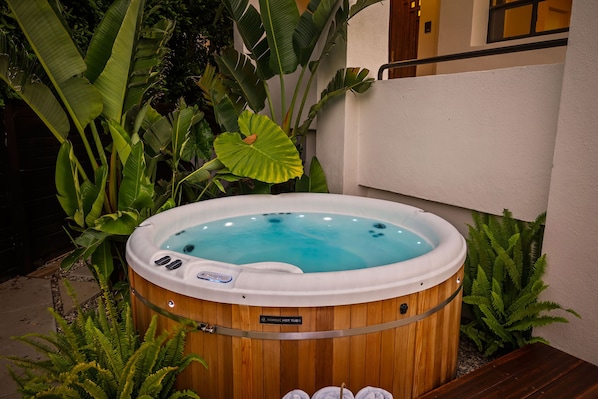 A hot tub with a wooden exterior, surrounded by lush greenery and a white wall.