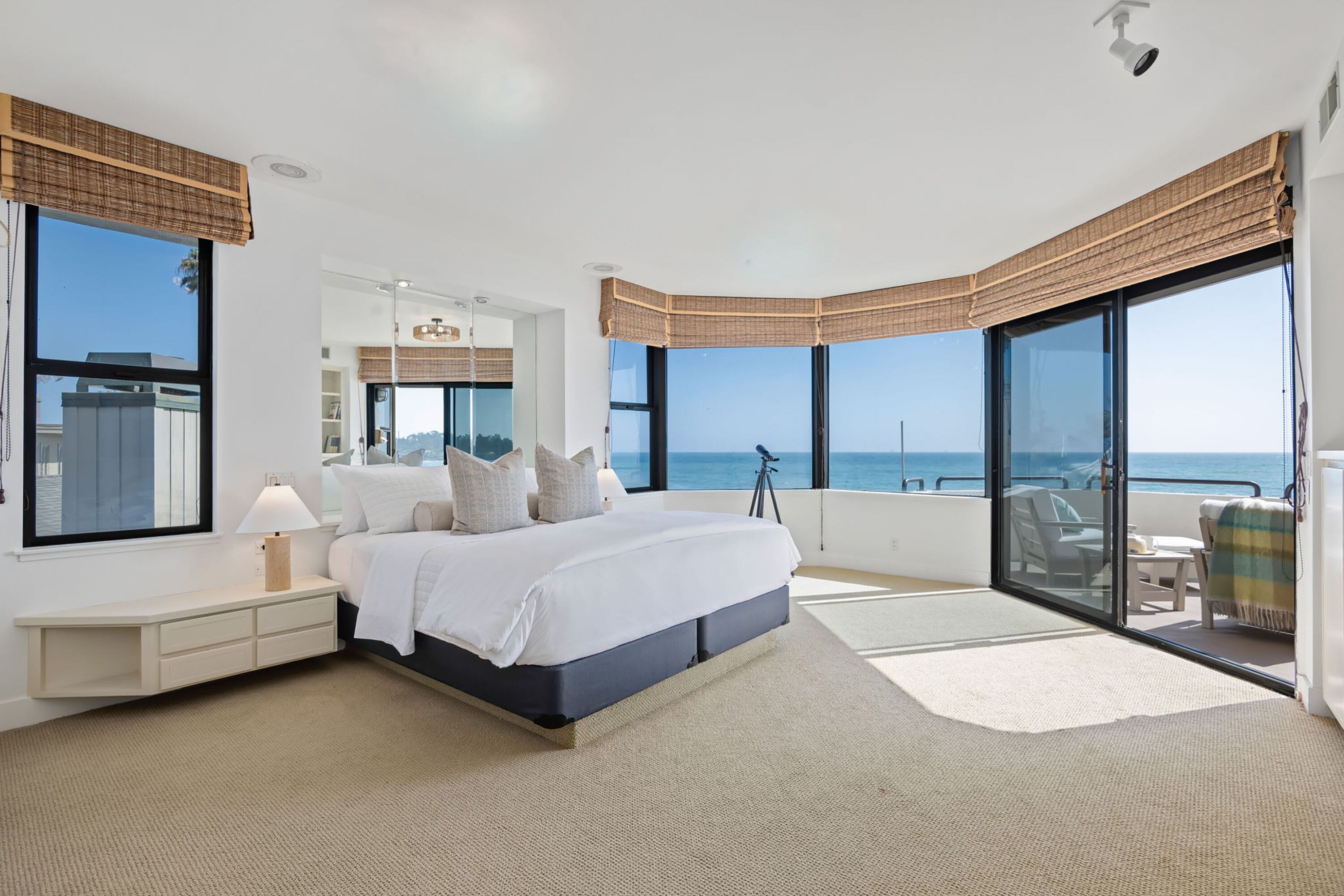 A bedroom with a large bed, a nightstand, a lamp, a telescope, and a view of the ocean.
