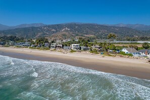 A coastal residential area with houses along the beach, a clear sky, and mountains in the background. - Exclusive Beachfront Estate | On the Sand, Hot Tub, EV Charger, Mtn & Sea Views (Carpinteria)