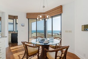 A dining area with a glass table and wicker chairs, a kitchen with a black refrigerator, and a view of the ocean through large glass doors. - Exclusive Beachfront Estate | On the Sand, Hot Tub, EV Charger, Mtn & Sea Views (Carpinteria)