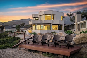 A modern multi-level building with large windows, a deck with chairs, and a mountainous backdrop. - Exclusive Beachfront Estate | On the Sand, Hot Tub, EV Charger, Mtn & Sea Views (Carpinteria)