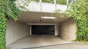A covered parking area with a concrete floor and walls, a closed garage door, and a height restriction sign. - Botanica Jelitkowo Morze i Slow (Gdańsk)