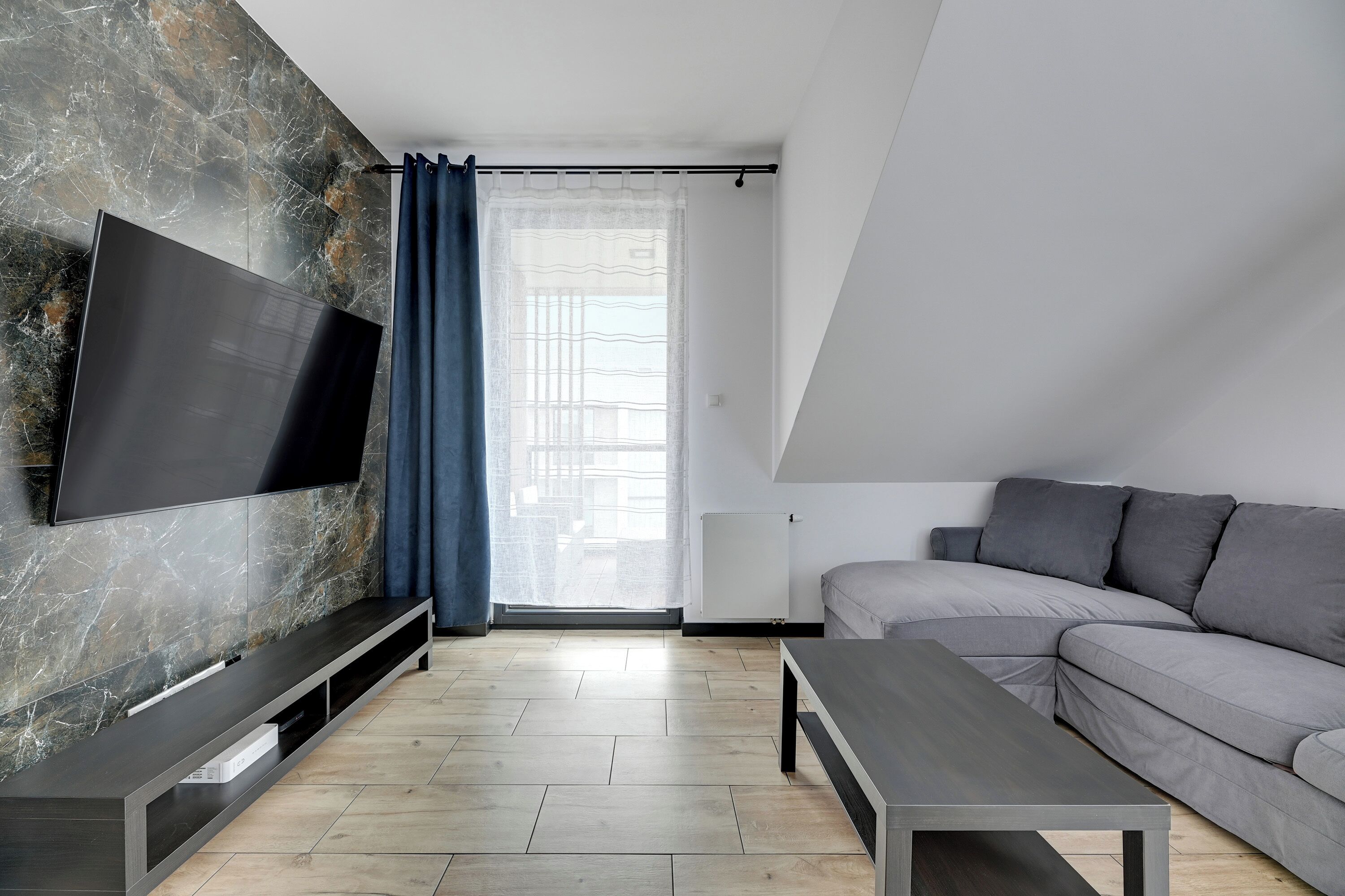 Ein modernes Wohnzimmer mit Flachbildfernseher an einer Marmorwand, einem grauen Sofa, einem Couchtisch und einem Fenster mit durchsichtigen Vorhängen.