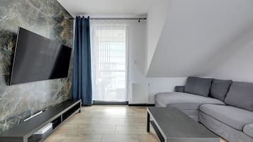 Ein modernes Wohnzimmer mit Flachbildfernseher an einer Marmorwand, einem grauen Sofa, einem Couchtisch und einem Fenster mit durchsichtigen Vorhängen.