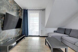A modern living room with a flat-screen TV mounted on a marble wall, a grey sofa, a coffee table, and a window with sheer curtains. - Botanica Jelitkowo Morze i Slow (Gdańsk)