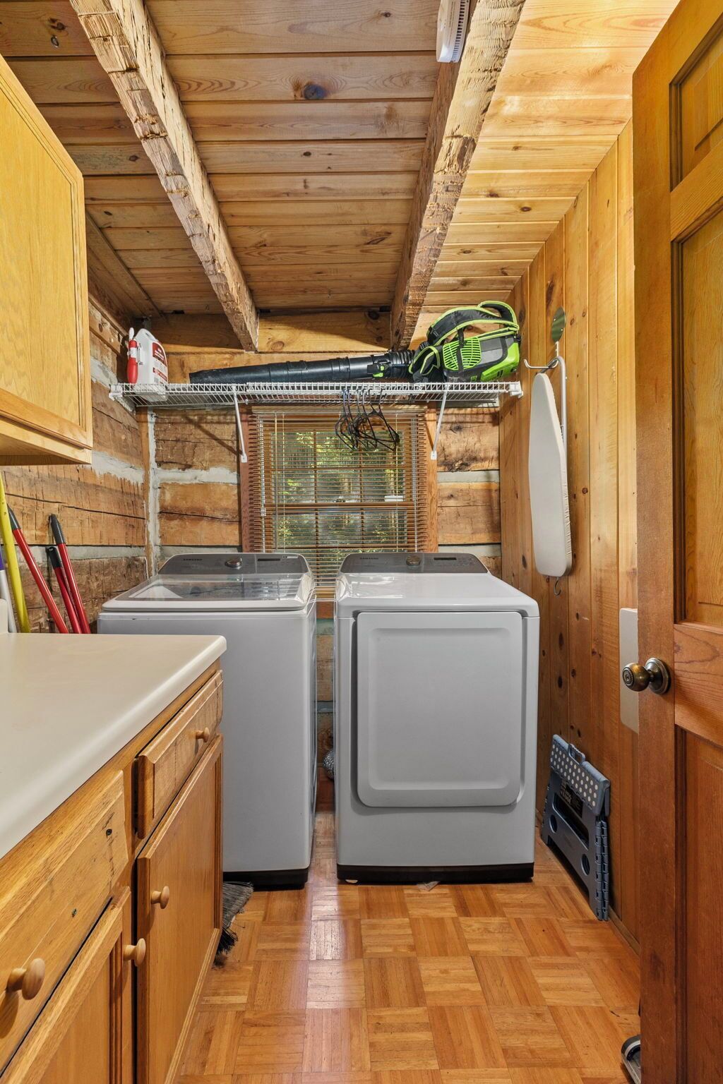 A laundry room with two washing machines, wooden cabinets, a shelf with cleaning supplies, and a door.