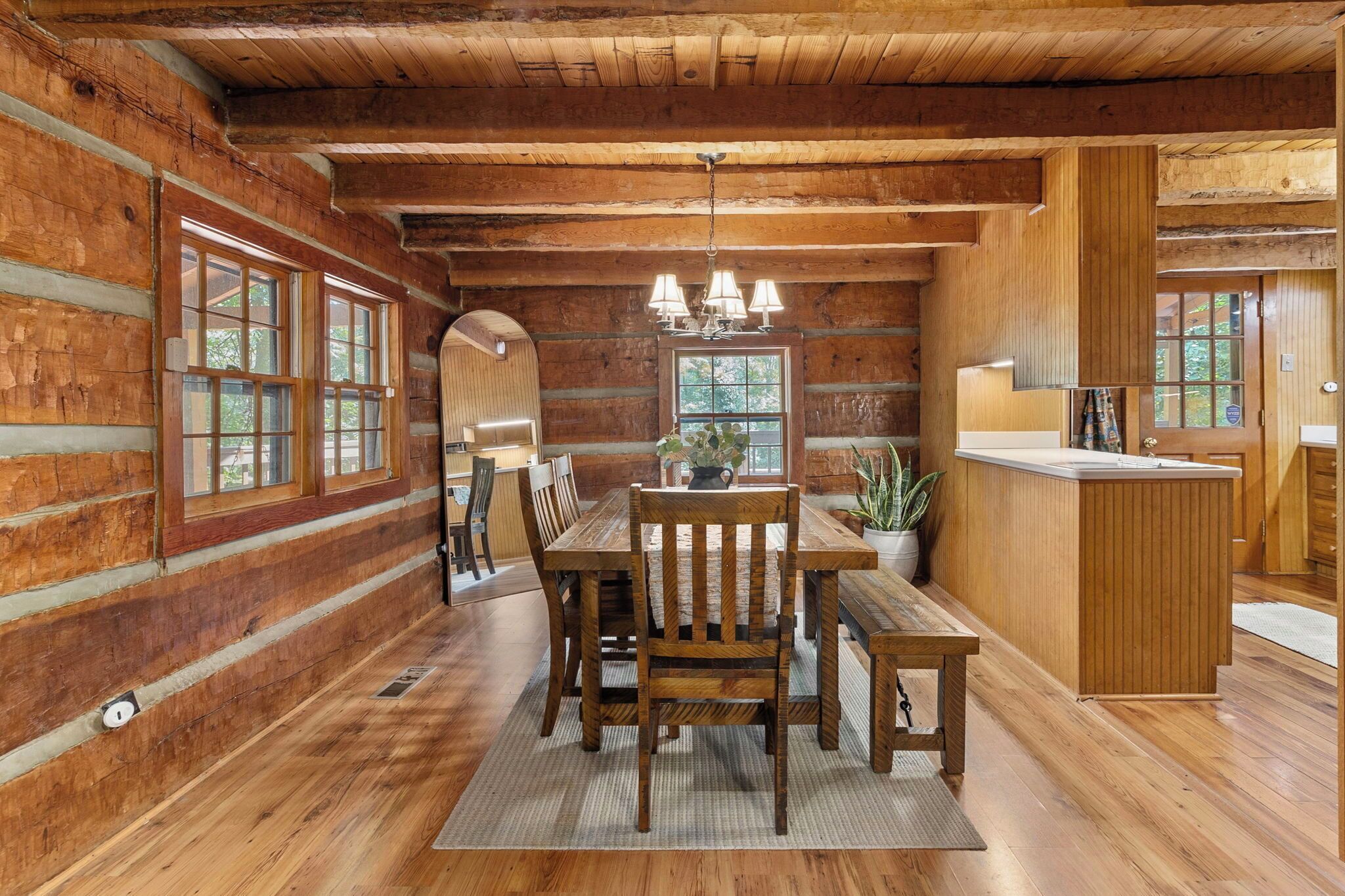 A wooden cabin interior with a dining area, kitchen, and large windows.