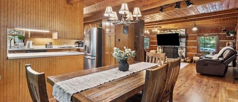 A wooden interior with a dining table, chairs, a kitchen area, and a living room with a fireplace.